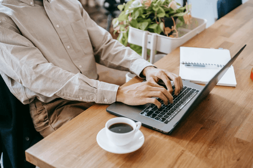 Een persoon zit aan een houten tafel te werken op een laptop, met een kop koffie en een notitieboekje naast zich.